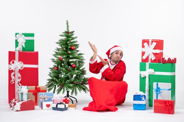 Surprised young adult dressed as Santa claus with gifts and decorated Christmas tree sitting on the ground pointing something on the right side on white background