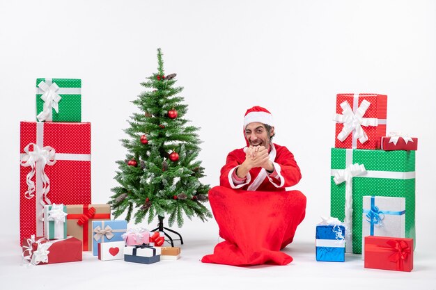 贈り物と白い背景の上の地面に座って飾られたクリスマスツリーでサンタクロースに扮した驚いた若い大人