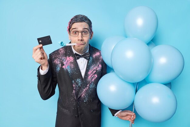 Удивленный молодой взрослый мужчина, одетый в праздничную одежду, дует в рог для вечеринки, держит кредитную карту, а куча надутых воздушных шаров празднует особый случай, изолированный на синем фоне Концепция праздника