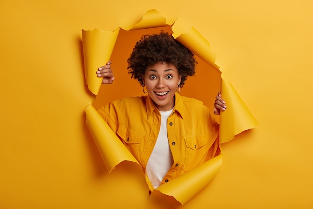 Surprised young African American woman stands in torn paper hole, dressed in stylish clothes, has excited cheerful expression