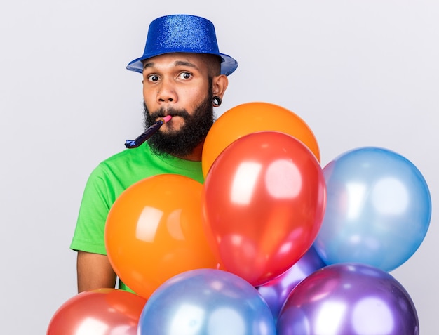 Sorpreso giovane ragazzo afroamericano che indossa un cappello da festa con in mano palloncini che soffiano fischietti di festa