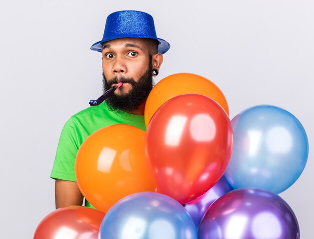 Surprised young afro-american guy wearing party hat holding balloons blowing party whistle 