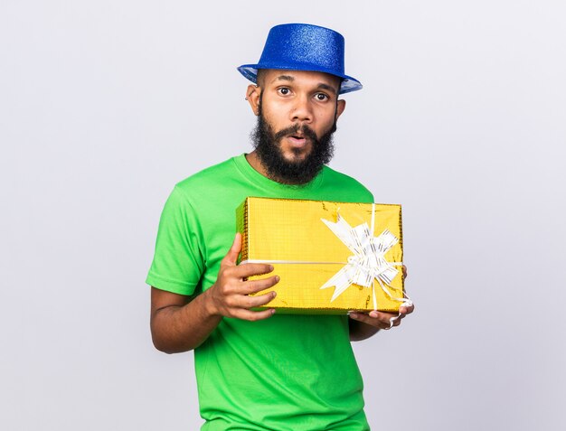 흰 벽에 격리된 선물 상자를 들고 파티 모자를 쓰고 놀란 젊은 아프리카계 미국인 남자