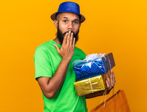 Surprised young afro-american guy wearing party hat holding gift boxes with bag covered mouth with hand 