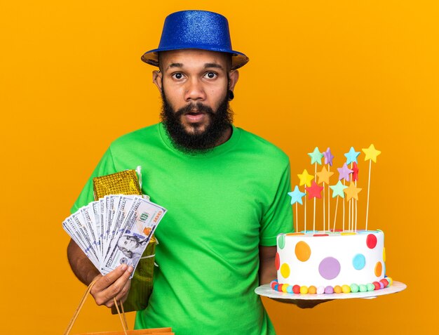 贈り物と現金でケーキを保持しているパーティーハットを身に着けている驚いた若いアフリカ系アメリカ人の男
