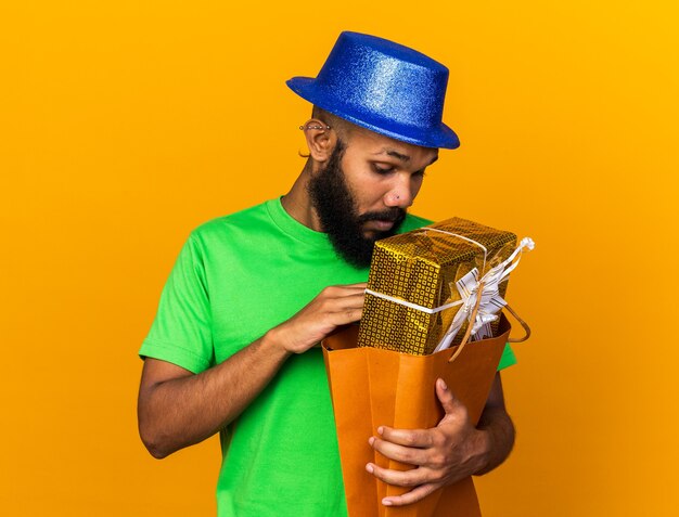 Surprised young afro-american guy wearing party hat holding and looking into gift bag 