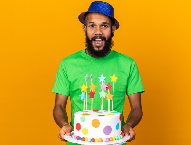 Surprised young afro-american guy wearing party hat holding out cake at camera 