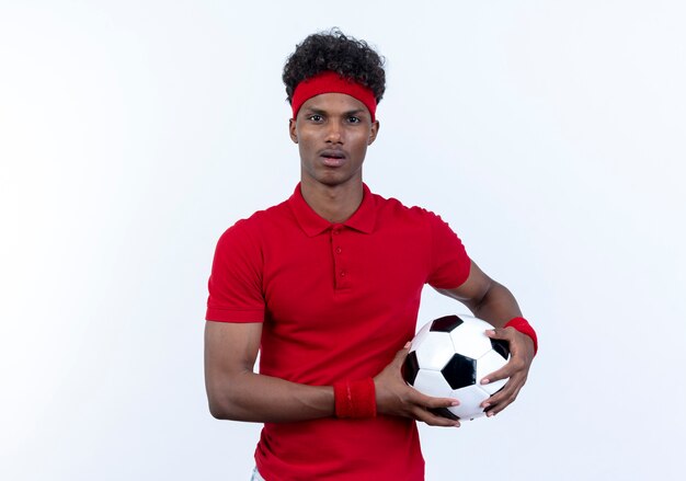 Surprised young afro-american sporty man wearing headband and wristband holding ball isolated on white