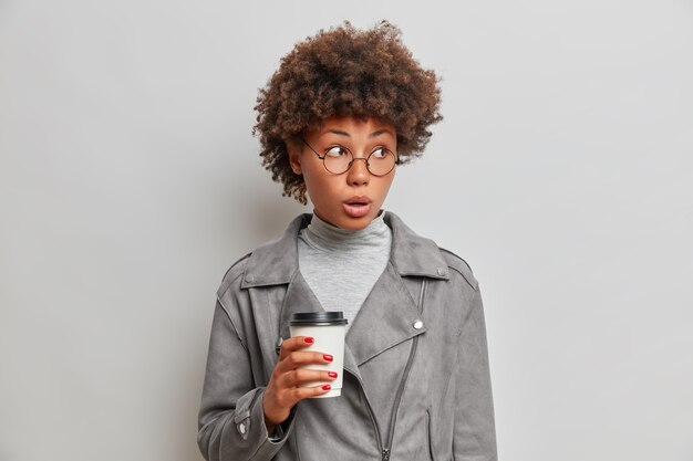 驚いた若いアフロの女性は、灰色のジャケットを着て、持ち帰り用のコーヒーを飲みます