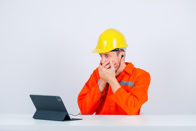 Surprised young architect is looking away by holding hands on mouth on white background