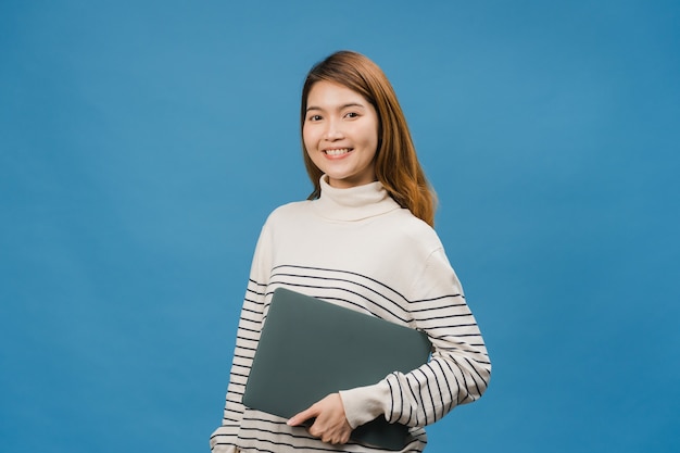Удивленная молодая азиатская леди держит ноутбук с позитивным выражением лица, широко улыбается, одетая в повседневную одежду и смотрит вперед на синей стене
