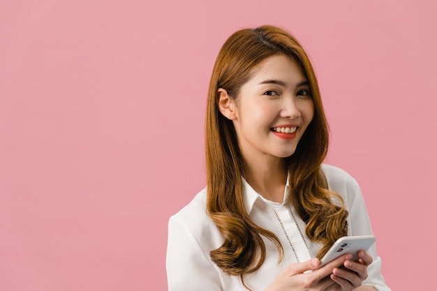 Surprised young Asia lady using mobile phone with positive expression, smiles broadly, dressed in casual clothing and looking at camera on pink background.