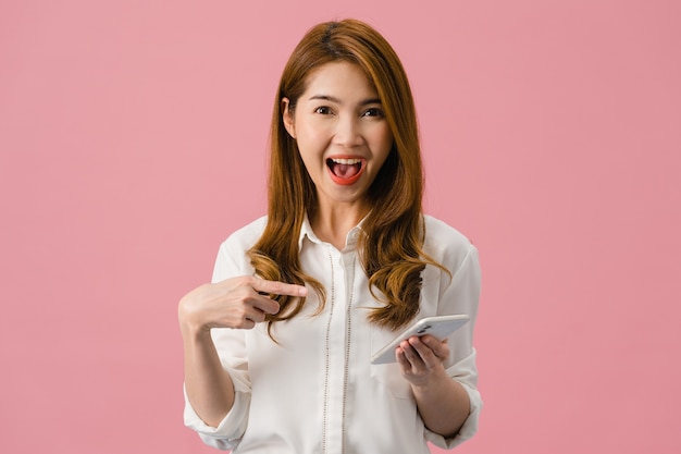 Удивленная молодая азиатская дама с позитивным выражением лица пользуется мобильным телефоном, широко улыбается, одета в повседневную одежду и смотрит в камеру на розовом фоне.