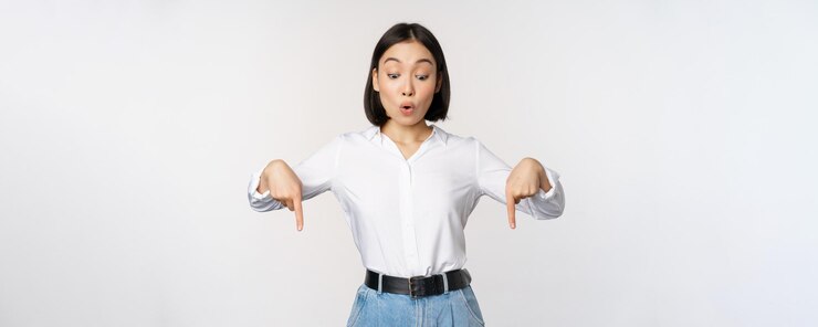 무료 Photo 놀란 젊은 아시아 여학생이 손가락을 아래로 가리키고 흰색 배경 위에 서 있는 놀란 표정으로 바라보고 있다