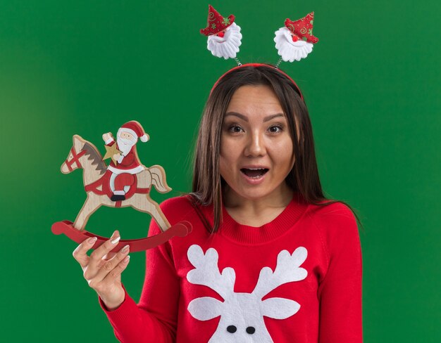 Surprised young asian girl wearing christmas hair hoop with sweater holding christmas toy isolated on green background