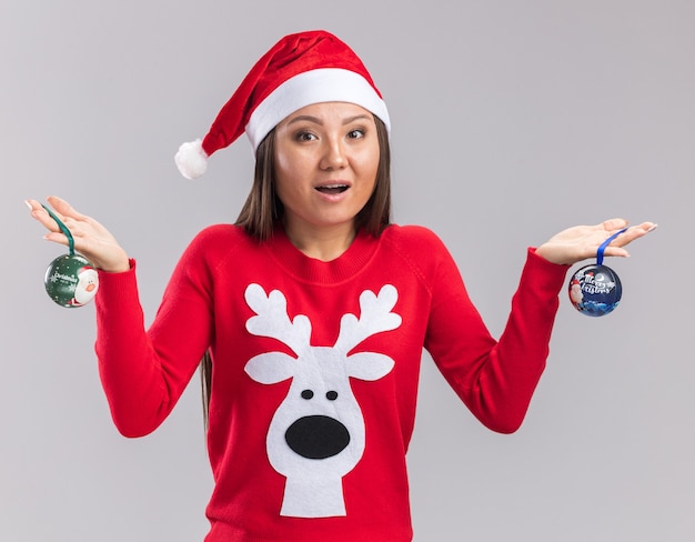 Surprised young asian girl wearing christmas hat with sweater holding christmas tree balls isolated on white background