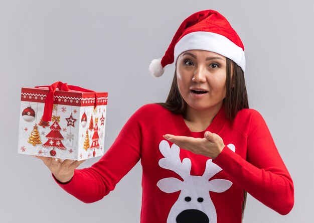 Free photo surprised young asian girl wearing christmas hat with sweater holding and points at gift box isolated on white wall