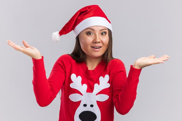 Surprised young asian girl wearing christmas hat with sweater spreading hands isolated on white wall