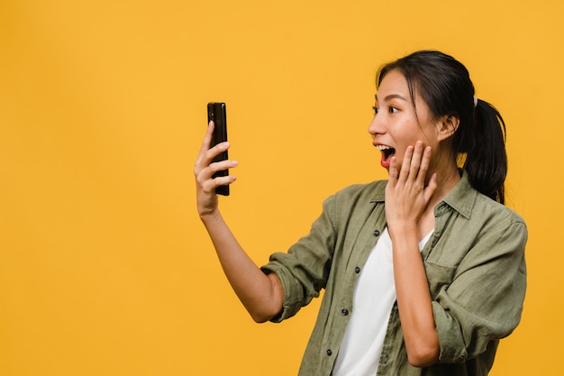 Surprised young Asian lady using mobile phone with positive expression, smiles broadly, dressed in casual clothing and stand isolated on yellow wall. Happy adorable glad woman rejoices success.