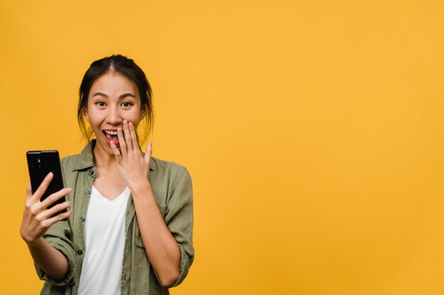 Surprised young Asian lady using mobile phone with positive expression, smiles broadly, dressed in casual clothing and stand isolated on yellow wall. Happy adorable glad woman rejoices success.