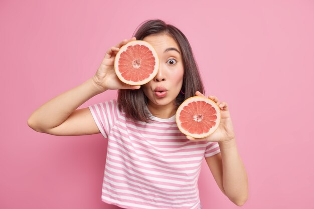 柑橘系の果物を屋内でポーズをとる驚いた若いアジア人女性は、新鮮なグレープフルーツの半分を保持し、健康的な栄養を保ち、ピンクの壁に隔離されたTシャツに身を包んだ表情に衝撃を与えました