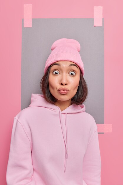 Surprised young Asian woman with healthy skin puckered lips stares at camera being impressed by something wears casual hoodie hat poses at studio with plastered sheet of paper for your promotion