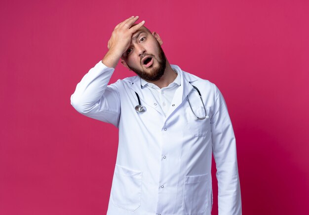 Surprised young bald male doctor wearing medical robe and stethoscope putting hand on forehead isolated on pink