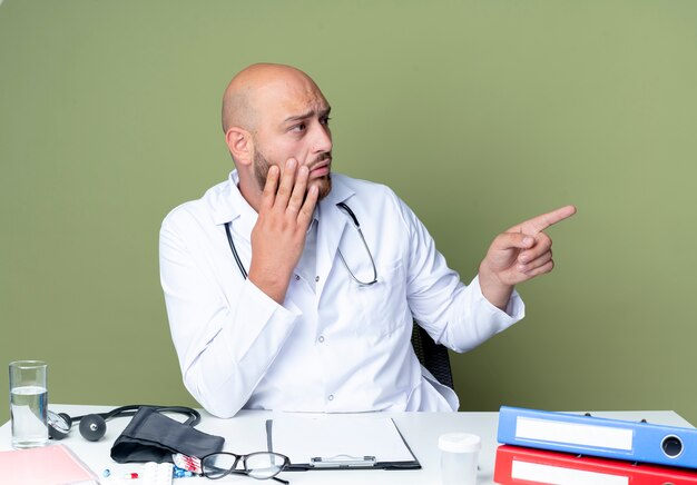 Surprised young bald male doctor wearing medical robe and stethoscope sitting at desk