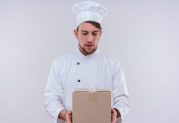 Удивленный молодой бородатый шеф-повар в белой форме держит коробку для доставки и смотрит на нее на белой стене