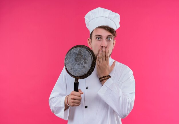 A surprised young bearded chef man in white uniform holding frying pan with hand on mouth while looking on a pink wall