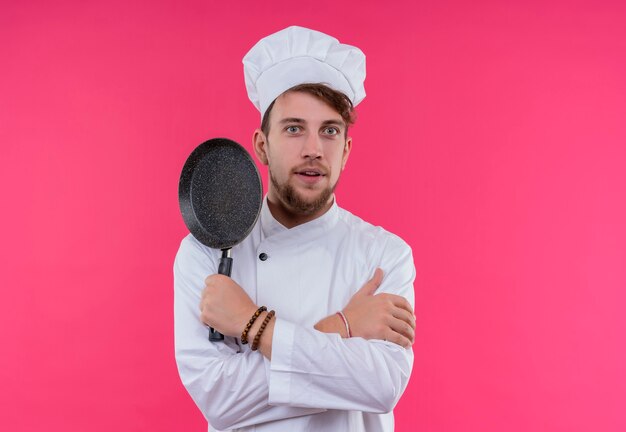 분홍색 벽에 찾고있는 동안 프라이팬을 들고 요리사 모자를 쓰고 흰색 제복을 입은 놀란 젊은 수염 요리사 남자