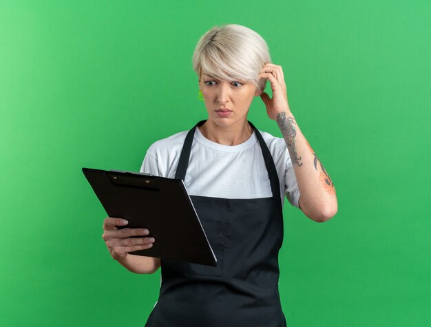 Surprised young beautiful female barber in uniform holding and looking at clipboard isolated on green background