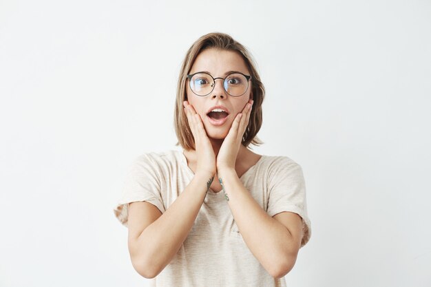 Surprised young beautiful girl in glasses with opened mouth and hands on cheeks 