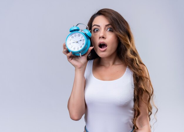 Surprised young beautiful girl holding alarm clock on isolated white wall with copy space