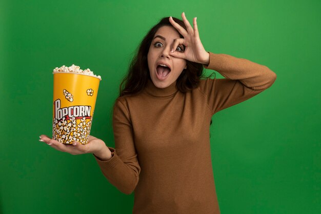 Surprised young beautiful girl holding bucket of popcorn showing look gesture isolated on green wall