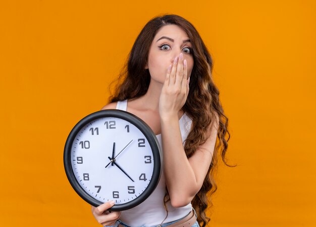 Surprised young beautiful girl holding clock with hand on mouth on isolated orange wall with copy space