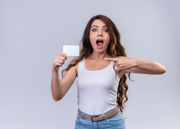 Surprised young beautiful girl holding credit card and pointing at it on isolated white wall with copy space