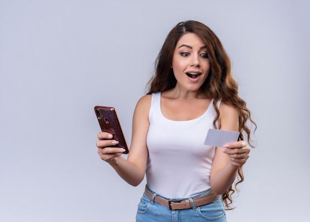 Удивленная молодая красивая девушка держит мобильный телефон и кредитную карту и смотрит на карту на изолированной белой стене с копией пространства
