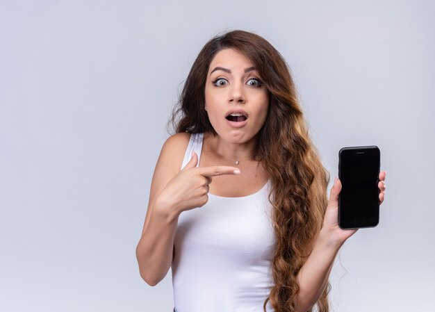 Surprised young beautiful girl holding mobile phone and pointing at it on isolated white wall with copy space