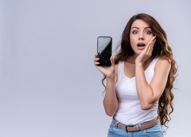 Surprised young beautiful girl showing mobile phone with hand on cheek on isolated white wall with copy space