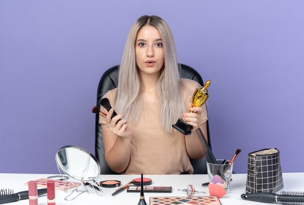 Surprised young beautiful girl sits at table with makeup tools holding winner cup with makeup brush isolated on blue background