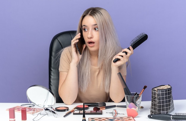Surprised young beautiful girl sits at table with makeup tools speaks on phone holding comb isolated on blue background