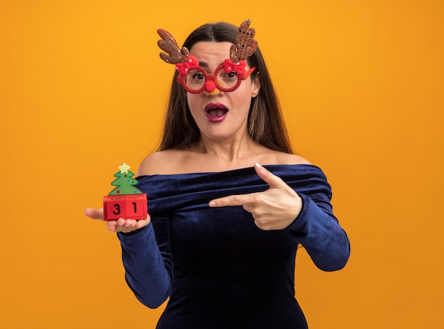 Surprised young beautiful girl wearing blue dress and christmas glasses holding and points at toy isolated on orange background