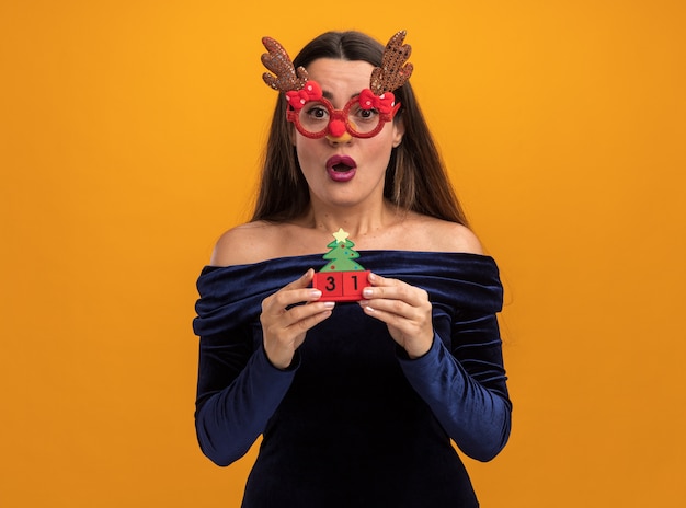 Foto gratuita giovane bella ragazza sorpresa che porta vestito blu e vetri di natale che tengono giocattolo isolato su fondo arancio