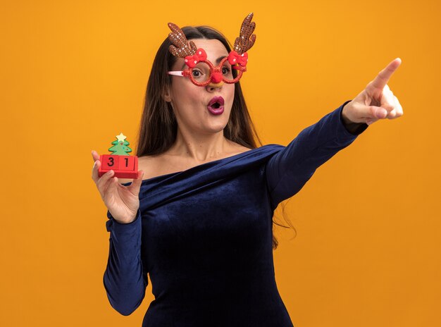 Surprised young beautiful girl wearing blue dress and christmas glasses holding toy points at side isolated on orange background