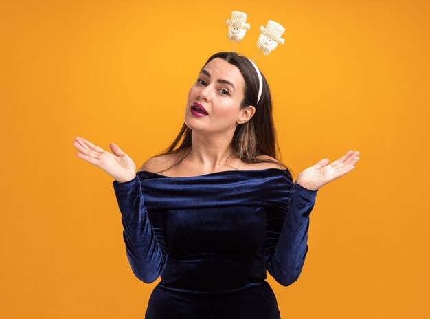 Surprised young beautiful girl wearing blue dress and christmas hair hoop spreading hands isolated on orange background