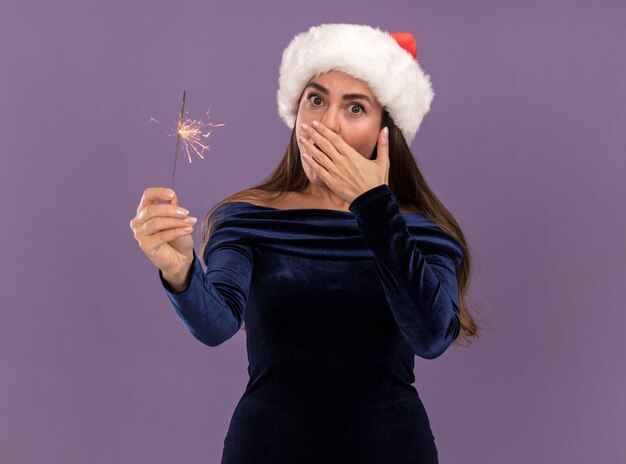 青いドレスと線香花火を保持しているクリスマス帽子を身に着けている驚いた若い美しい少女は、紫色の背景で隔離の手で口を覆った
