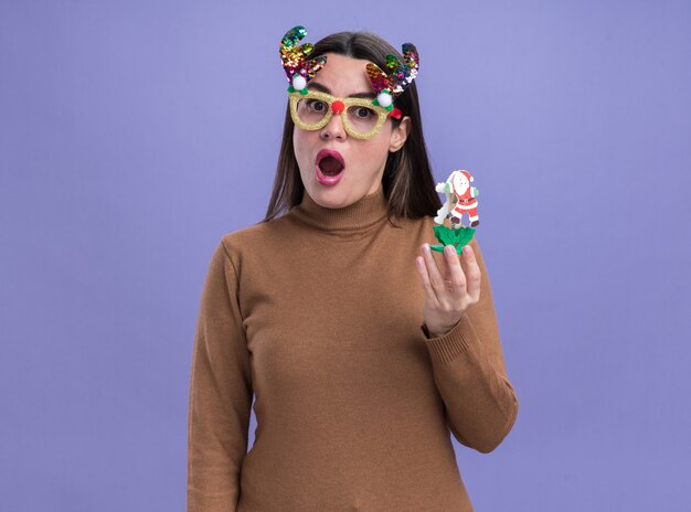 Удивленная молодая красивая девушка в коричневом свитере с рождественскими очками держит игрушку на синем фоне