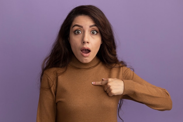 Surprised young beautiful girl wearing brown turtleneck sweater points at herself isolated on purple wall