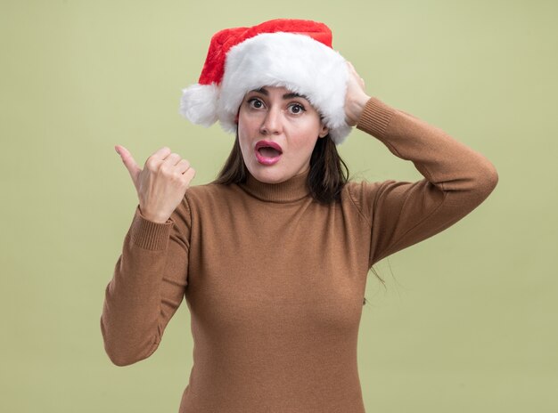Surprised young beautiful girl wearing christmas hat points at side isolated on olive green background with copy space